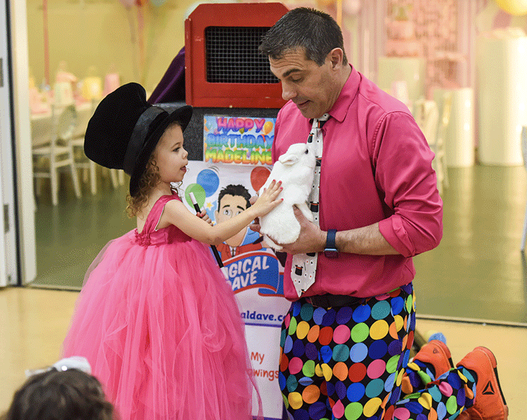 magical-dave-nyc-magic-shows-kids3 Magical Dave in a colorful polka-dot outfit showing his live white rabbit to a young girl in a pink dress at a birthday party